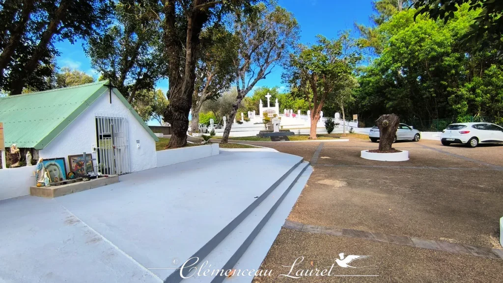 Petite chapelle ou oratoire blanc au toit en tôle verte à gauche de l'image, au Calvaire de Petite-Île. Devant l'oratoire, des statues religieuses de la Vierge Marie et de saint Joseph, ainsi que des cadres pieux, sont déposés en offrande sur un socle. Des marches en béton mènent à une grande esplanade propre, ouvrant sur le chemin de croix et le calvaire blanc en arrière-plan entouré d'arbres. - Photo : Clémenceau Lauret (Licence CC BY-SA 4.0)