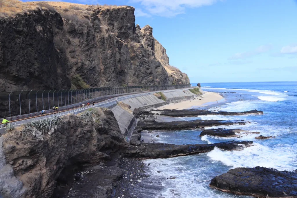 Vue panoramique sur la falaise volcanique du Cap La Houssaye à Saint-Paul, la route littorale avec des cyclistes et les rochers noirs battus par l'écume des vagues. Photo : Clémenceau Lauret (CC BY-SA 4.0)