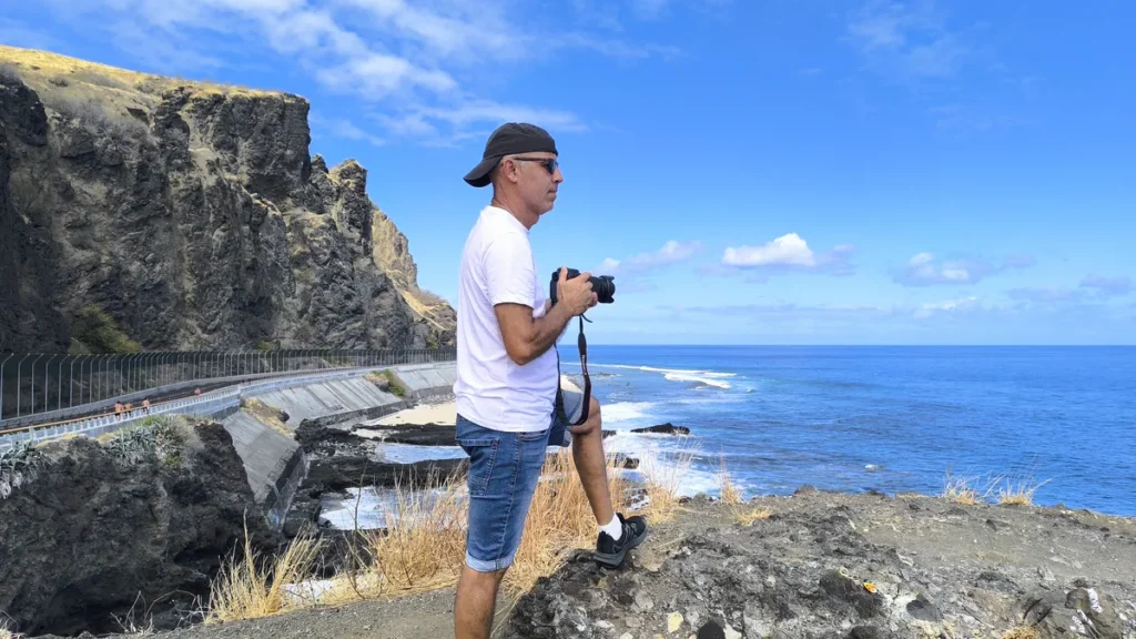 Clémenceau Lauret debout en profil sur un éperon rocheux du Cap La Houssaye à Saint-Paul, casquette et appareil photo en main devant la falaise et la mer. Photo : Clémenceau Lauret (CC BY-SA 4.0)
