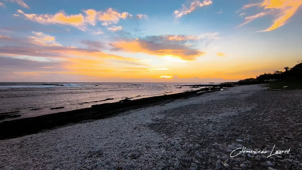 Vue panoramique au format paysage sur la plage de débris coralliens et de basalte de Grand-Bois à Saint-Pierre, baignée par les teintes dorées et orangées du coucher de soleil avec un ciel parsemé de nuages blonds, photographiée par Clémenceau Lauret (CC BY-SA 4.0).