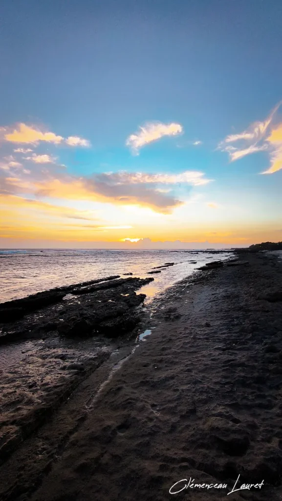 Cliché vertical cadrant la grève rocheuse et le sable sombre de la côte de Grand-Bois à Saint-Pierre, révélant la réfraction de la lumière du soleil couchant sur l'océan Indien et les petites retenues d'eau, réalisé par Clémenceau Lauret (CC BY-SA 4.0).