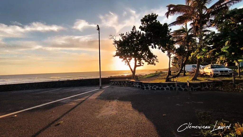 Promenade aménagée du littoral de Grand-Bois à Saint-Pierre avec des arbres en contre-jour, des véhicules stationnés et la mer scintillante sous une douce lumière d'un fin de journée, cliché de Clémenceau Lauret (CC BY-SA 4.0).