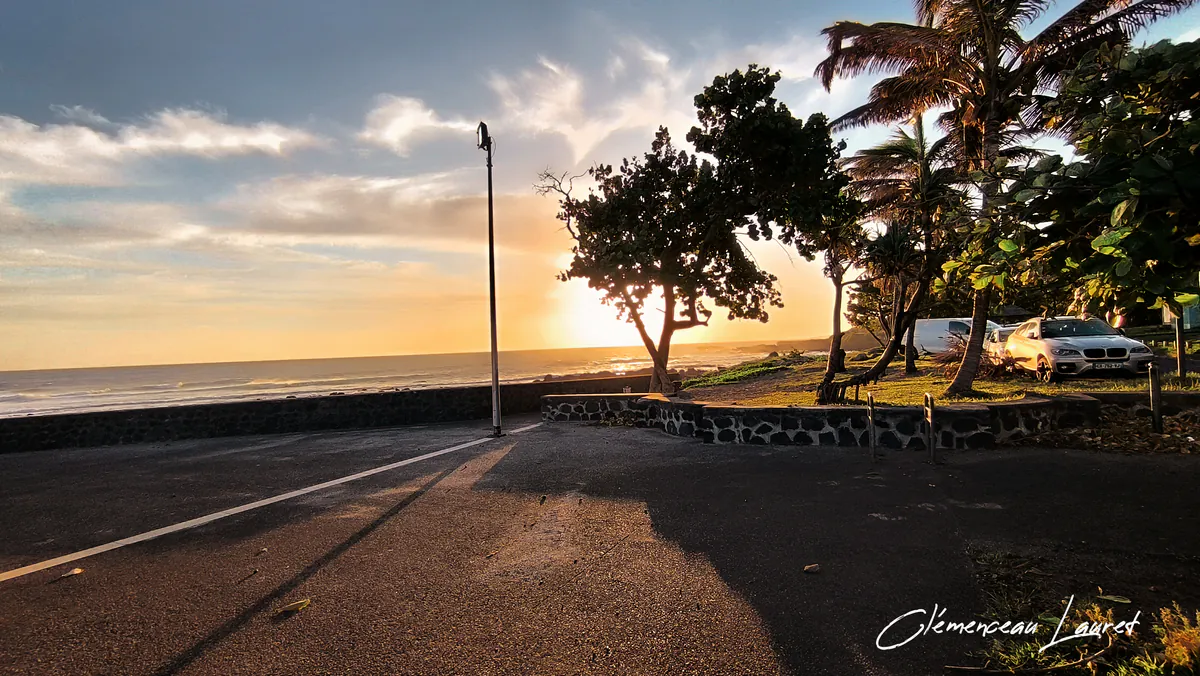 Promenade aménagée du littoral de Grand-Bois à Saint-Pierre avec des arbres en contre-jour, des véhicules stationnés et la mer scintillante sous une douce lumière d'un fin de journée, cliché de Clémenceau Lauret (CC BY-SA 4.0).