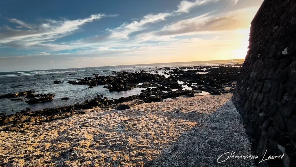 Coin de plage de sable et de corail à Grand-Bois, Saint-Pierre, bordé d'un mur de soutènement en roches taillées, avec une mer calme parsemée de rochers basaltiques au soleil couchant, photographié par Clémenceau Lauret (CC BY-SA 4.0).