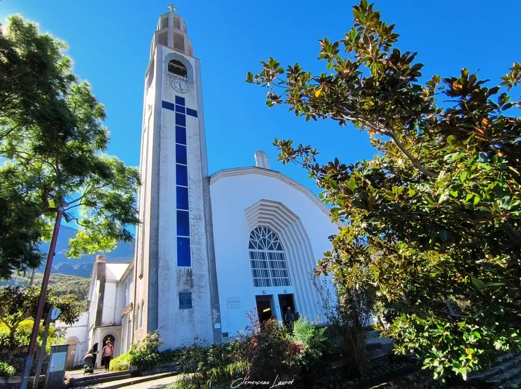 Photographie en contre-plongée mettant en valeur l'architecture moderniste de la façade et du clocher de l'église Notre-Dame-des-Neiges à Cilaos, entourée d'arbres verdoyants sous un ciel pur et ensoleillé, réalisée par Clémenceau Lauret.
