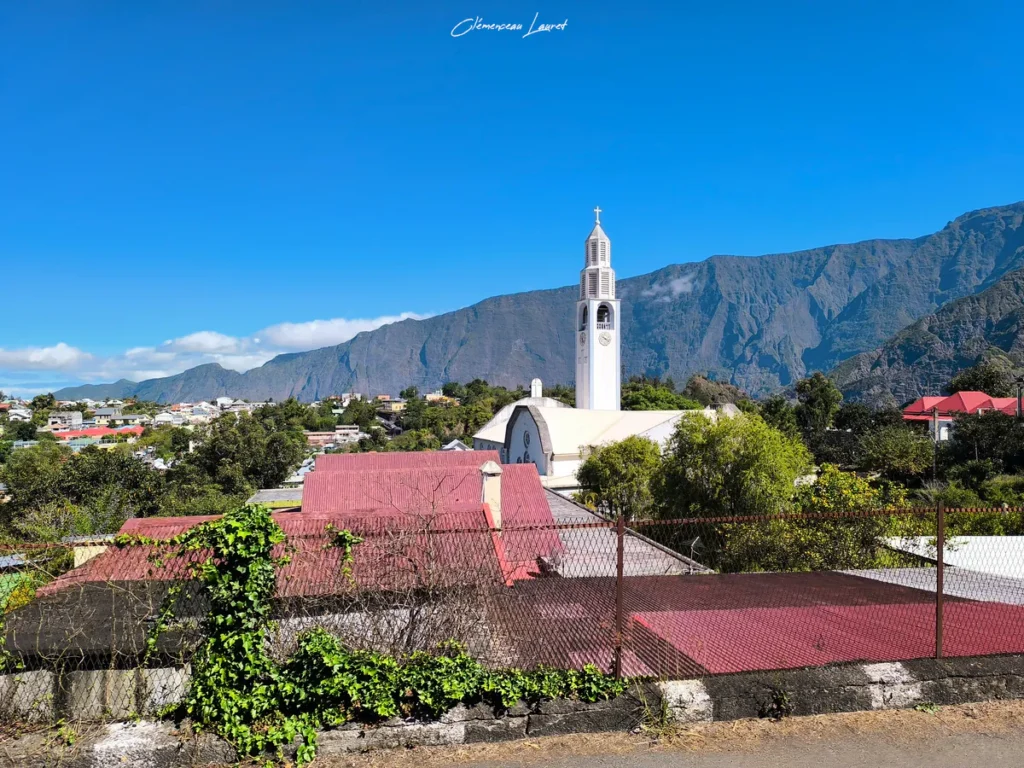 Photographie en plongée capturant le clocher blanc de l'église Notre-Dame-des-Neiges s'élevant au-dessus des toits colorés du village de Cilaos, entouré par l'imposante muraille minérale des montagnes du cirque, réalisée par Clémenceau Lauret.