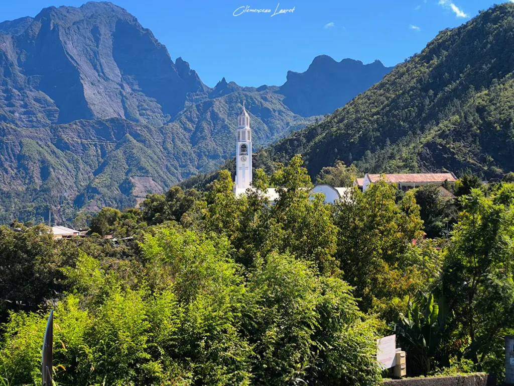 Photographie panoramique montrant une vue distante de l'église Notre-Dame-des-Neiges émergeant d'une végétation tropicale luxuriante, avec les sommets escarpés et majestueux du cirque de Cilaos se découpant sous un ciel bleu éclatant, réalisée par Clémenceau Lauret.