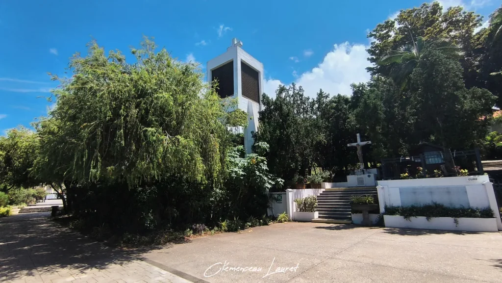 Clocher moderne blanc et calvaire entourés d'arbres tropicaux aux Avirons à La Réunion sous un ciel bleu partiellement nuageux.