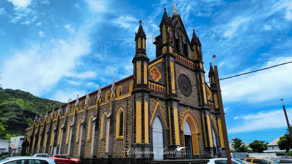 Vue extérieure de la façade principale et du flanc de l'église Notre-Dame de la Délivrance sous un ciel bleu nuageux, montrant son architecture néogothique aux pierres basaltiques et teintes jaunes, à Saint-Denis, La Réunion — Photo : Clémenceau Lauret (CC BY-SA 4.0).