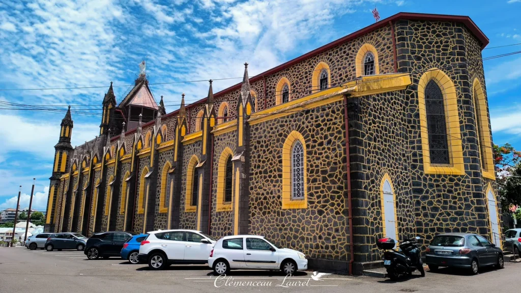 Façade latérale et toiture inclinée de l'église Notre-Dame de la Délivrance bordée par un parking, au cœur de Saint-Denis, La Réunion — Photo : Clémenceau Lauret (CC BY-SA 4.0).