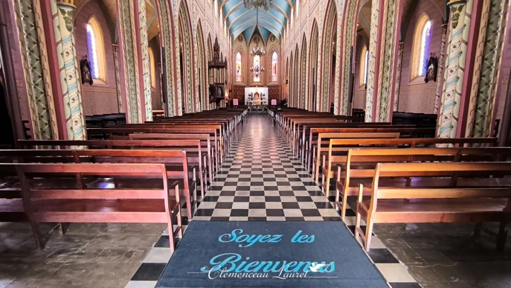 Allée centrale de la nef de l'église Notre-Dame de la Délivrance menant vers l'autel, encadrée par des bancs en bois et des colonnes peintes, à Saint-Denis, La Réunion — Photo : Clémenceau Lauret (CC BY-SA 4.0).