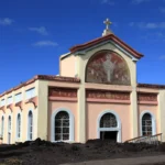 Angle de trois-quarts de l'église Notre-Dame des Laves émergeant des blocs de lave à Piton Sainte-Rose sous un ciel bleu.