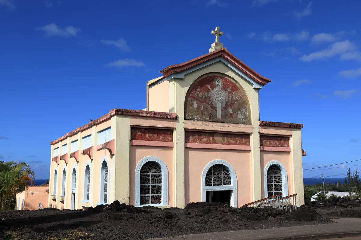 Angle de trois-quarts de l'église Notre-Dame des Laves émergeant des blocs de lave à Piton Sainte-Rose sous un ciel bleu.