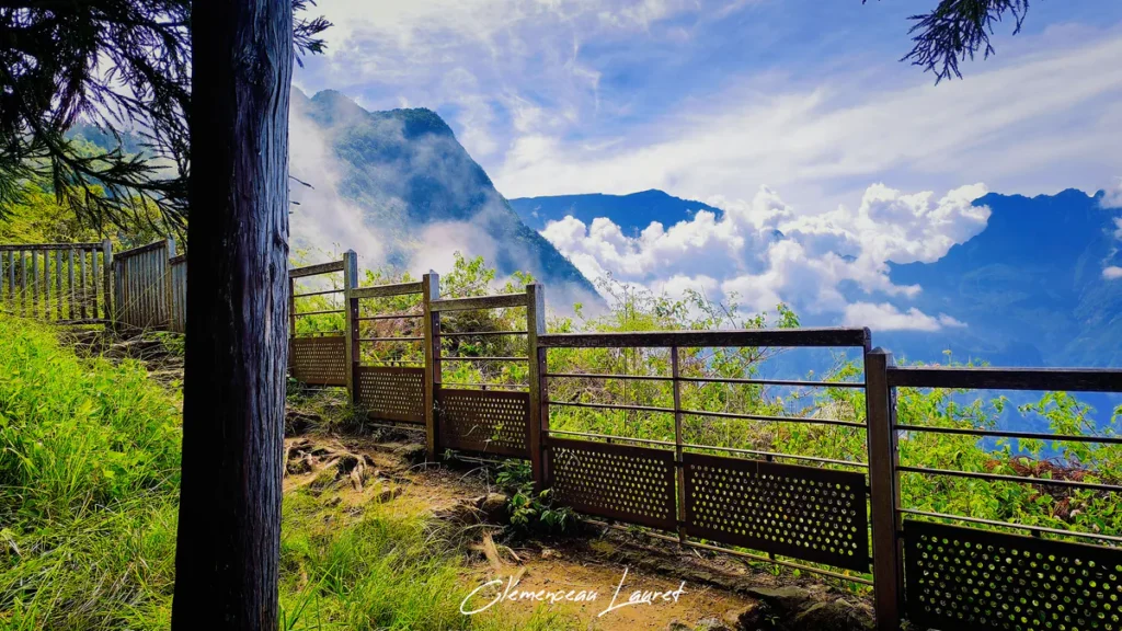 Vue latérale d'un sentier de terre bordé d'une clôture en bois et métal à La Fenêtre des Makes, Saint-Louis, avec un premier plan végétalisé et une vue imprenable sur les montagnes et nuages de Cilaos.
