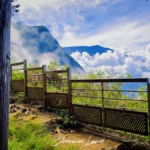 Vue latérale d'un sentier de terre bordé d'une clôture en bois et métal à La Fenêtre des Makes, Saint-Louis, avec un premier plan végétalisé et une vue imprenable sur les montagnes et nuages de Cilaos.