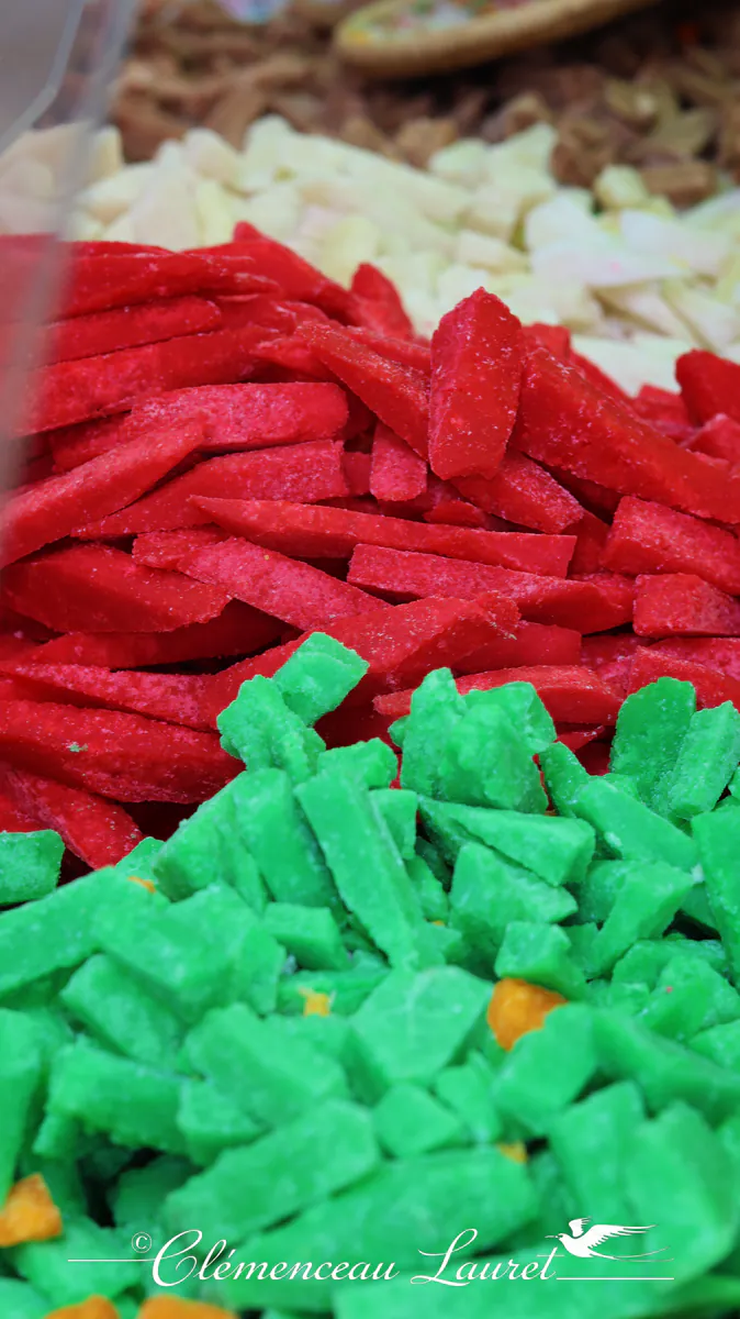 Cadre vertical immersif montrant un étal gourmand de confiseries traditionnelles réunionnaises. Le bas du cliché met en avant des fondantes vertes empilées, suivies d'une rangée de fondantes rouges intenses au centre, et de douceurs blanches et dorées en arrière-plan flou. Ambiance de marché local capturée avec une grande précision de texture et de contraste. Photo : Clémenceau Lauret à Le Tampon (Licence CC BY-SA 4.0).