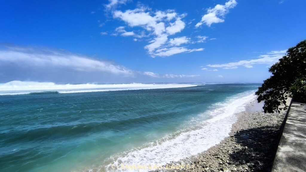 Vue panoramique depuis la côte montrant l'eau turquoise se transformant en écume blanche sur la plage de galets de Saint-Pierre, avec au large une large bande de vagues puissantes générées par la houle australe - Photo : Clémenceau Lauret (Licence CC BY-SA 4.0)
