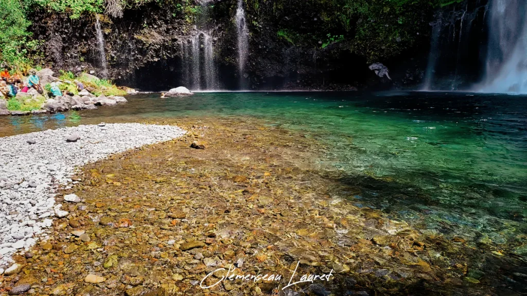 Rive de galets clairs et fond d'eau cristalline sous une falaise moussue à Grand Bassin, La Réunion.