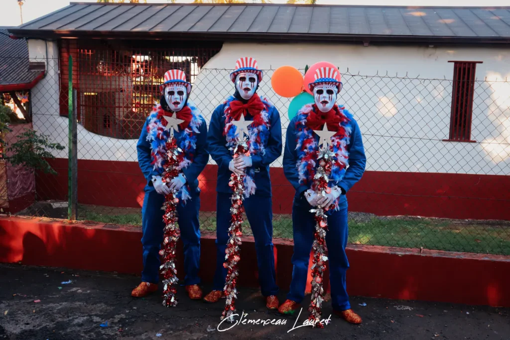 Trois carnavaliers en tenue bleue et masques blancs peints défilent au carnaval Karmon à Saint-Louis, tenant des bâtons ornés de guirlandes et d'étoiles sous une belle lumière.