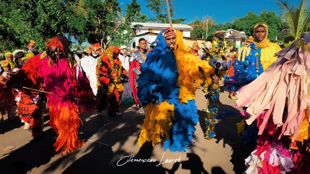 Cortège dynamique du Karmon à Saint-Louis montrant plusieurs participants recouverts de franges textiles multicolores en mouvement sur l'asphalte ensoleillé.