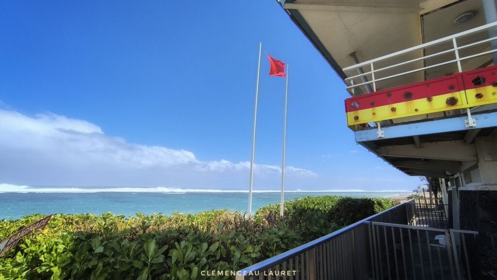Gros plan en contre-plongée sur le drapeau rouge flottant à côté du bâtiment du poste de secours de Saint-Pierre, surplombant la mer déchaînée par la houle australe sous un ciel ensoleillé - Photo : Clémenceau Lauret (Licence CC BY-SA 4.0)