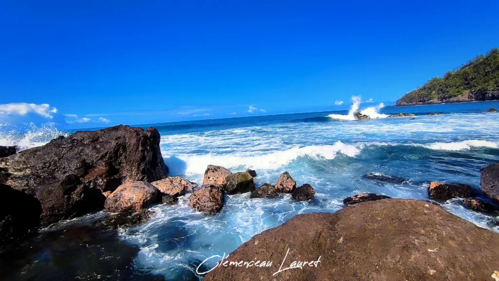 Écume des vagues déferlant sur les rochers basaltiques de la baie de Manapany à Saint-Joseph sous un grand ciel bleu.