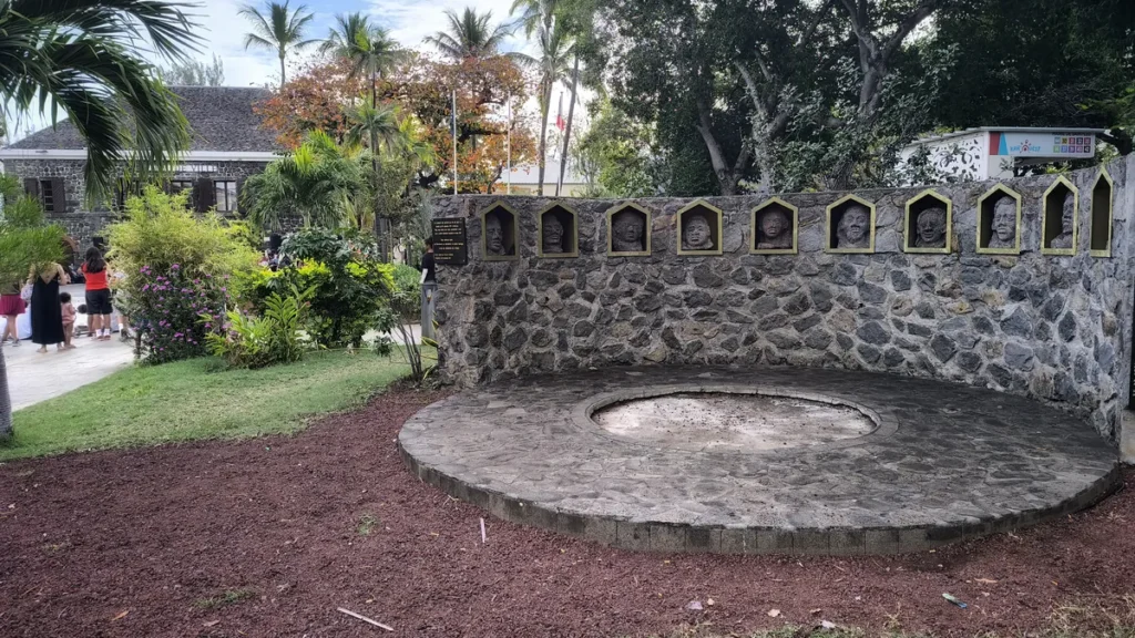 Structure demi-circulaire en maçonnerie de pierre de basalte du mémorial de la révolte des esclaves de 1811 avec des visages sculptés intégrés dans des niches dorées, située dans le parc de la mairie à Saint-Leu. Photo : Clémenceau Lauret (CC BY-SA 4.0).