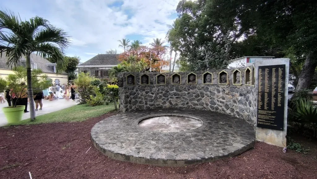 Vue d'ensemble du monument commémoratif de la révolte des esclaves de 1811 à Saint-Leu, présentant un muret arqué en pierres taillées, des sculptures faciales et une plaque stèle portant les noms des insurgés. Photo : Clémenceau Lauret (CC BY-SA 4.0).