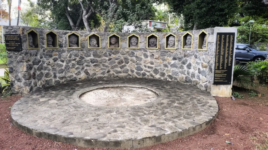 Vue frontale de l'œuvre monumentale en pierres sombres et niches en arc avec têtes sculptées honorant les insurgés de 1811 sur le site Mémorial de la Révolte des Esclaves de Saint-Leu. Photo : Clémenceau Lauret (CC BY-SA 4.0).