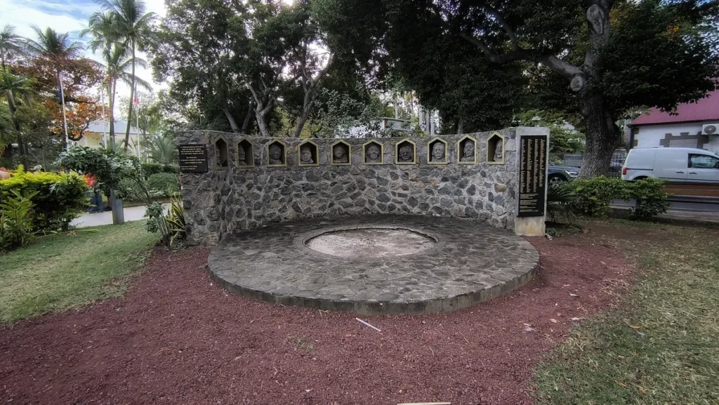 Angle élargi du mémorial circulaire de la révolte des esclaves de 1811 au cœur du jardin communal de Saint-Leu, entouré d'arbres tropicaux et de végétation luxuriante. Photo : Clémenceau Lauret (CC BY-SA 4.0).
