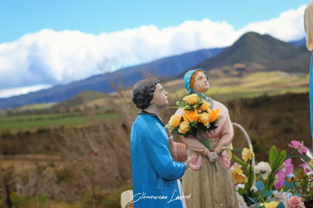 Statues de deux petits bergers en prière tenant des fleurs au premier plan de l'oratoire Notre-Dame des Fougères à la Plaine des Cafres, avec en arrière-plan le paysage verdoyant et les montagnes de La Réunion.