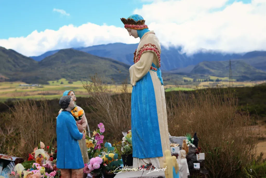 Statue colorée de la Vierge Marie surplombant les bergers et des bouquets de fleurs à l'oratoire de Notre-Dame des Fougères, face au panorama des remparts et montagnes de la Plaine des Cafres.