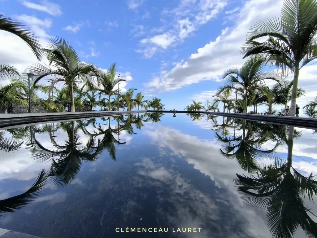 Miroir d'eau rectangulaire reflétant parfaitement le ciel nuageux et la rangée de palmiers au Parc des Palmiers à Le Tampon, Île de La Réunion. Photo : Clémenceau Lauret (CC BY-SA 4.0).