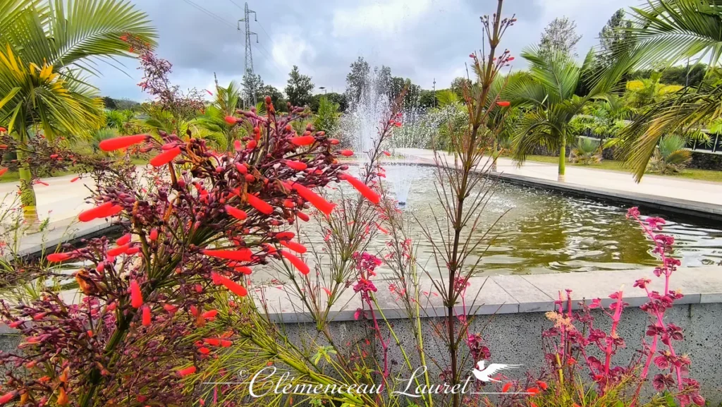 Fleurs tropicales rouges et orangées au premier plan devant un bassin animé par plusieurs jets d'eau au Parc des Palmiers à Le Tampon, Île de La Réunion. Photo : Clémenceau Lauret (CC BY-SA 4.0).