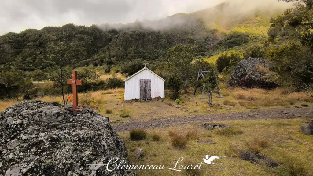 Croix en bois sur roche basaltique face à la chapelle de Marla sur un terrain herbeux entouré de montagnes à La Réunion.