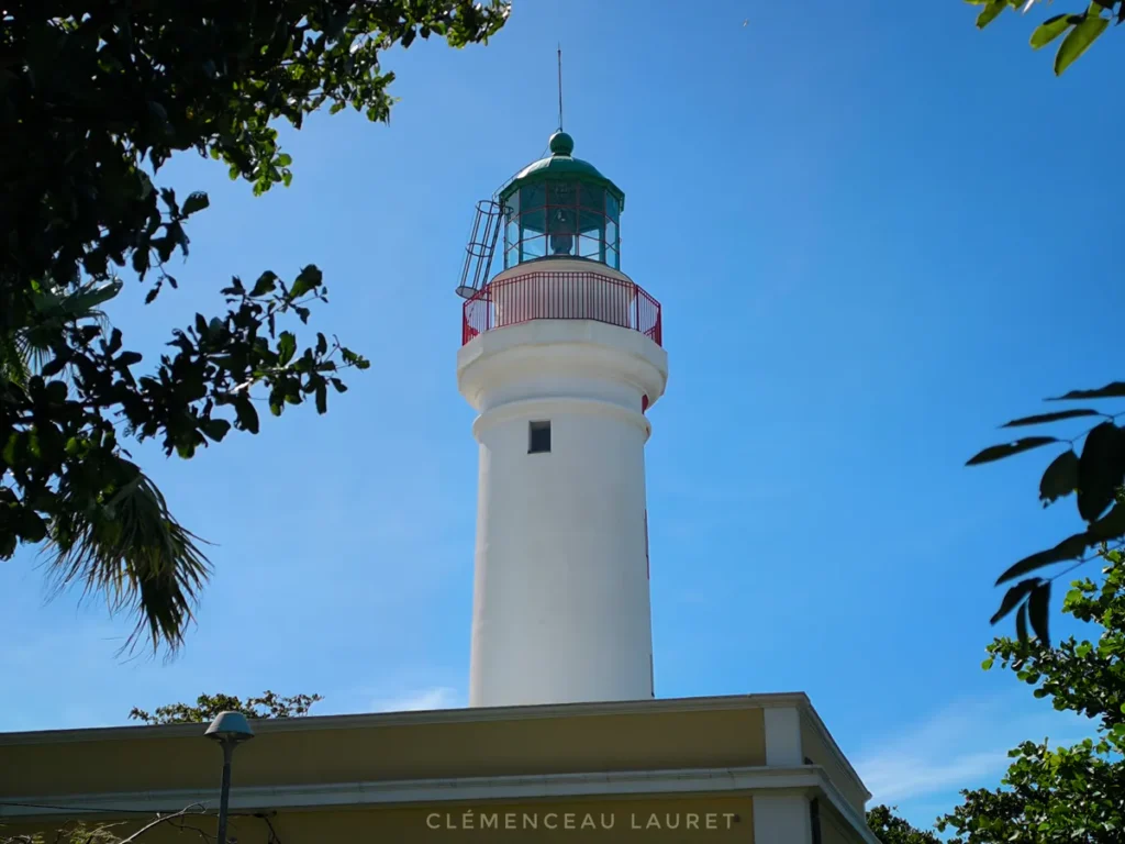 Vue rapprochée de la tour blanche du Phare de Bel-Air à Sainte-Suzanne sous un ciel bleu radieux, entourée par le feuillage vert d'arbres tropicaux. Au sommet, la lanterne verte et son garde-corps rouge se détachent fièrement au-dessus du bâtiment d'accueil ocre jaune. Photographie prise par Clémenceau Lauret (CC BY-SA 4.0).