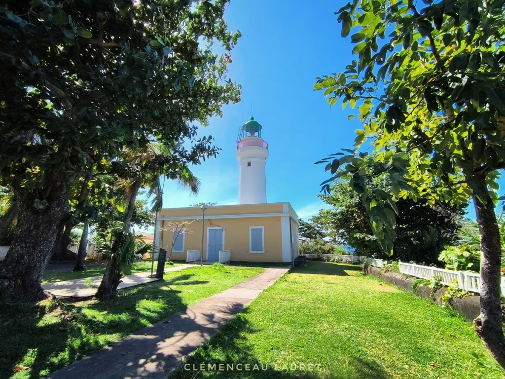 Vue panoramique de l'édifice du Phare de Bel-Air à Sainte-Suzanne, entouré d'un jardin gazonné très vert et bordé de grands arbres tropicaux. Un chemin pavé mène à la bâtisse ocre jaune dominée par sa majestueuse tour blanche et son dôme vert, sous un soleil éclatant. Photographie prise par Clémenceau Lauret (CC BY-SA 4.0).