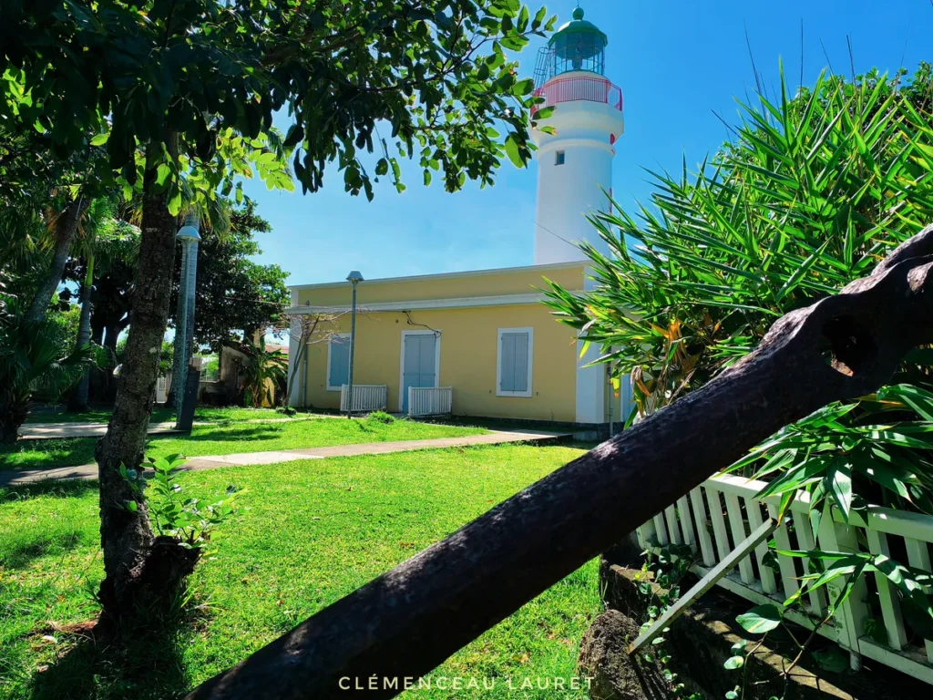 Perspectives sur le domaine du Phare de Bel-Air situé sur le littoral de Sainte-Suzanne, encadré par des arbres tropicaux, une végétation luxuriante et un ancien élément métallique de mouillage au premier plan. La tour du phare blanche à lanterne verte s'élève au-dessus du bâtiment principal. Photographie prise par Clémenceau Lauret (CC BY-SA 4.0).