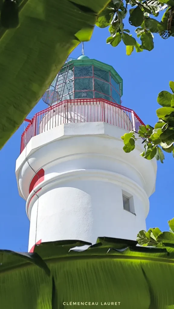Prise de vue en contre-plongée spectaculaire sur la lanterne verte et le garde-corps rouge du Phare de Bel-Air à Sainte-Suzanne, cadrée à travers les larges palmes vertes d'un bananier. Le sommet du phare se détache vivement sous un ciel azur intense. Photographie prise par Clémenceau Lauret (CC BY-SA 4.0).