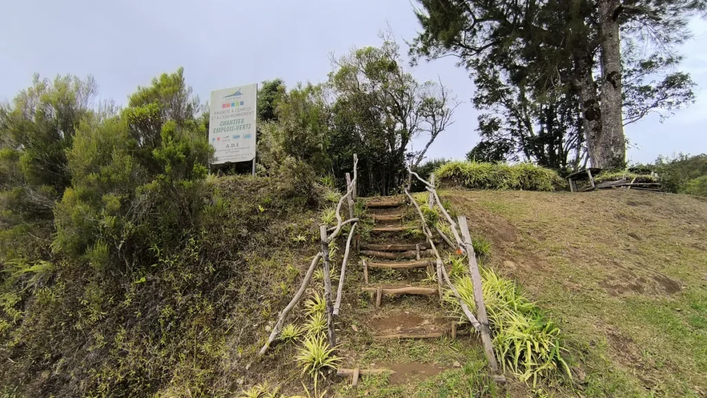 Escalier rustique en bois aménagé à même la terre pour accéder aux hauts de l'aire de pique-nique de Piton Fougères à Sainte-Marie, entouré d'une végétation verdoyante et surmonté d'un panneau du chantier d'insertion Emplois Verts A.D.E — Photo : Clémenceau Lauret (CC BY-SA 4.0).