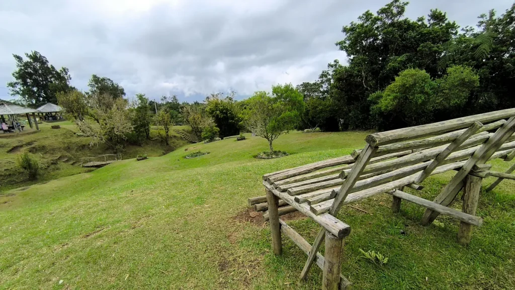 Grand banc en bois brut orienté vers un paysage vallonné et verdoyant, offrant un espace de repos privilégié sur l'aire de pique-nique aménagée de Piton Fougères à Sainte-Marie, avec des kiosques et une passerelle au loin — Photo : Clémenceau Lauret (CC BY-SA 4.0).