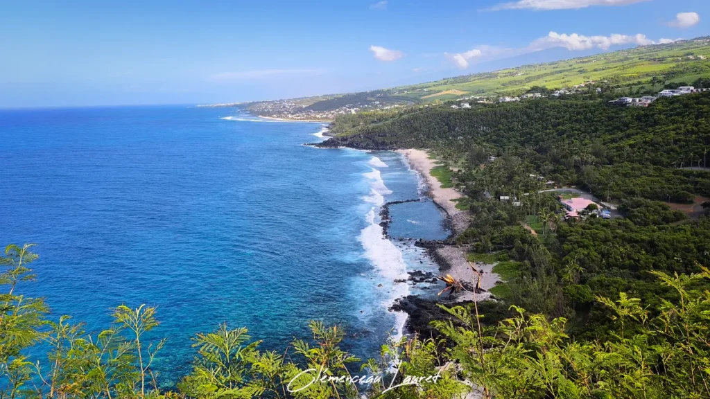 Vue panoramique de la plage de Grande Anse et son bassin de baignade depuis les hauteurs verdoyantes du piton à Petite-Île sous un ciel bleu clair.