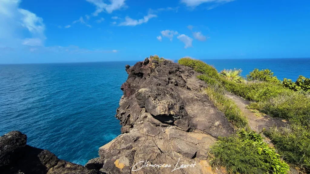 Éperon rocheux en volcanisme sombre bordé de végétation tropicale surplombant les eaux turquoises à Petite-Île sous un ciel ensoleillé.