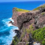 Cap volcanique teinté de rouge et de basalte noir dominant la mer bleue avec des branches de filaos au premier plan à Petite-Île.