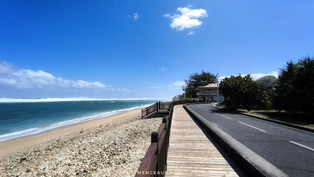 Vue panoramique de la promenade en lattes de bois le long du littoral de Saint-Pierre sous un ciel bleu dégagé, avec les vagues imposantes de la forte houle de septembre 2026 déferlant sur la plage de sable et de corail - Photo : Clémenceau Lauret (Licence CC BY-SA 4.0)