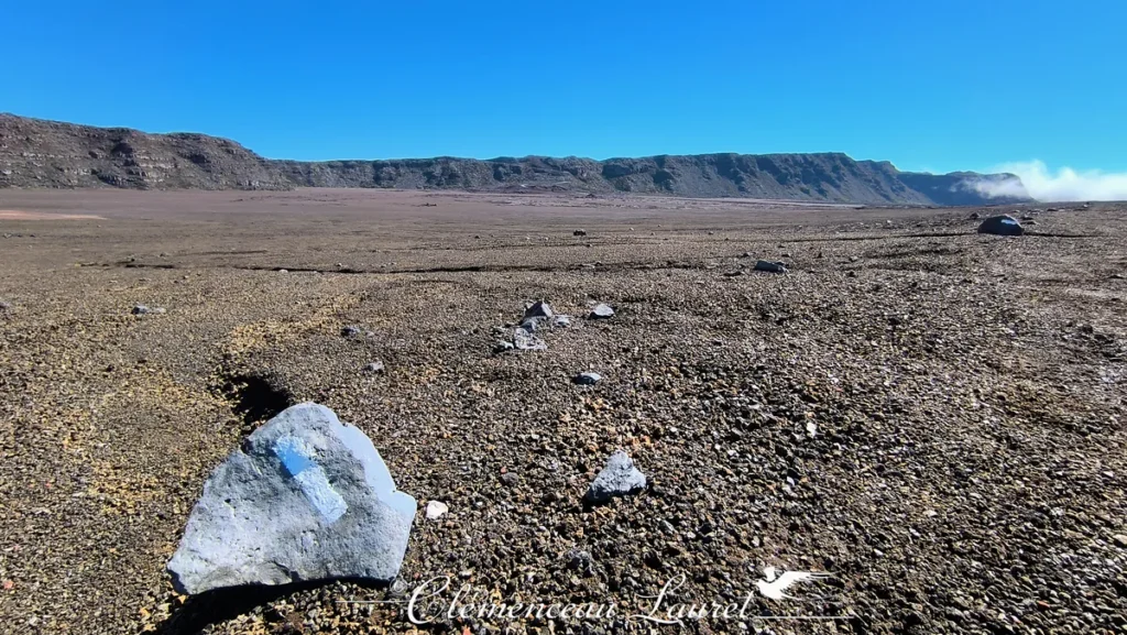 Vue panoramique de la Plaine des Sables sous un ciel bleu éclatant, mettant en valeur un sentier de randonnée balisé par une pierre marquée au premier plan et les falaises volcaniques en arrière-plan à La Réunion, Clémenceau Lauret, CC BY-SA 4.0.