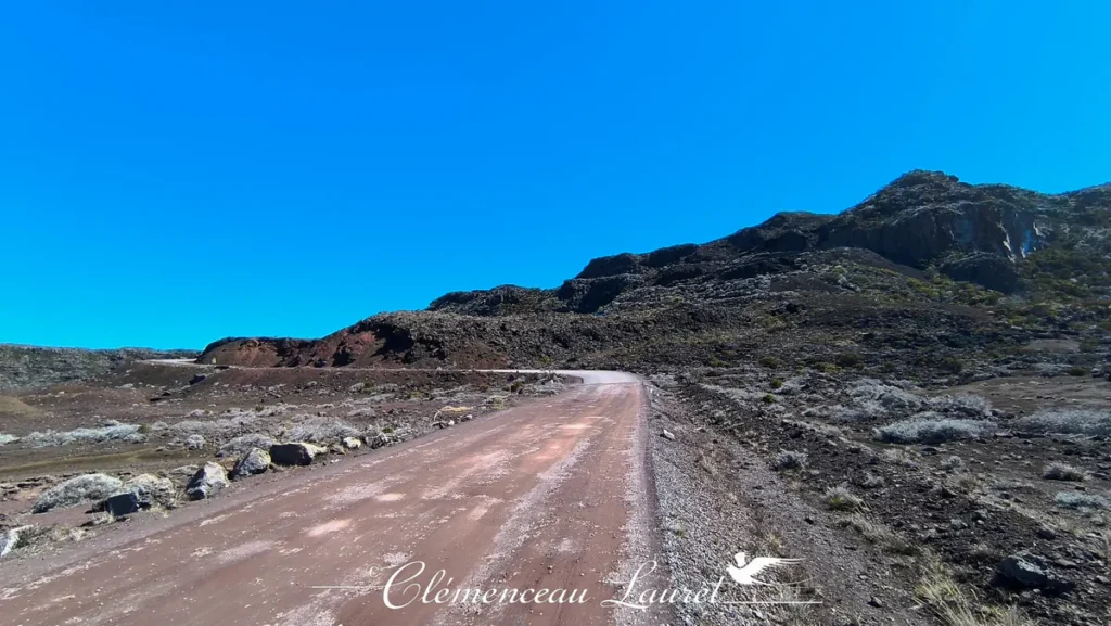 Piste sinueuse bordée de roches volcaniques et de végétation basse s'enfonçant dans les paysages de la Plaine des Sables à La Réunion, Clémenceau Lauret, CC BY-SA 4.0.