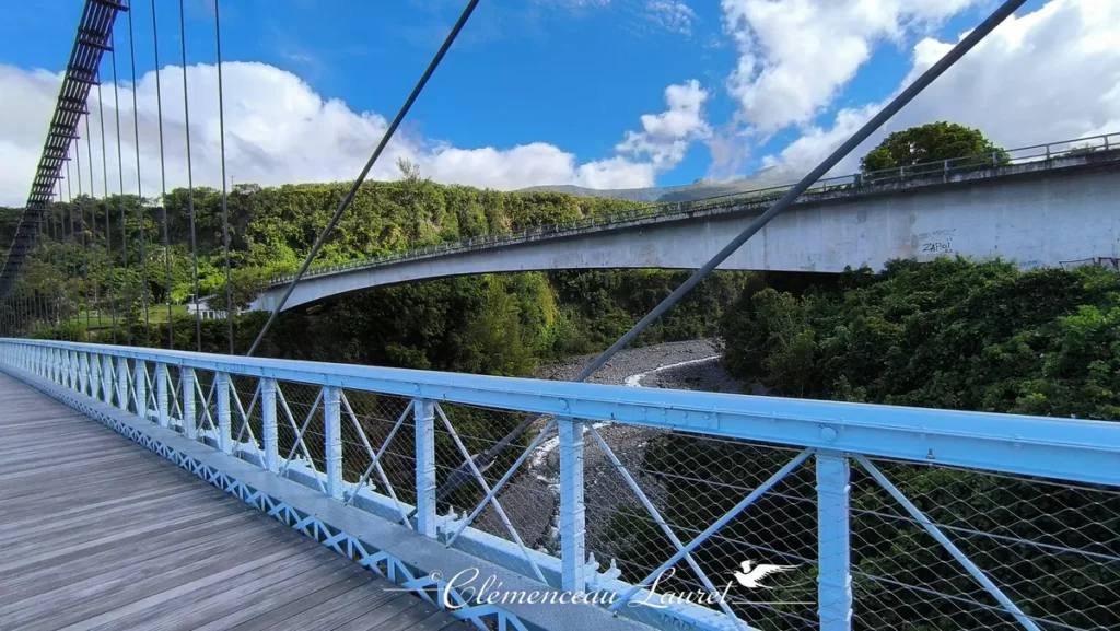 Prise de vue latérale depuis la rambarde métallique bleu ciel du pont suspendu, révélant la ravine en contrebas et le pont routier en béton moderne en arrière-plan. Le lit de la rivière est composé de galets sombres et d'un mince filet d'eau. La végétation verdoyante recouvre les berges escarpées sous un ciel bleu parsemé de nuages volumineux. - Photo : Clémenceau Lauret (Licence CC BY-SA 4.0)