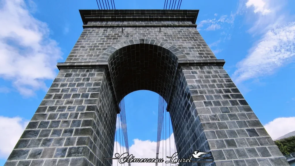 Vue en contre-plongée spectaculaire de la monumentale pile en maçonnerie de pierre taillée du pont suspendu. L'arche centrale en voûte s'élève vers un ciel bleu clair, encadrée par des câbles de suspension en acier tendus qui s'élèvent vers le sommet de la structure. - Photo : Clémenceau Lauret (Licence CC BY-SA 4.0)