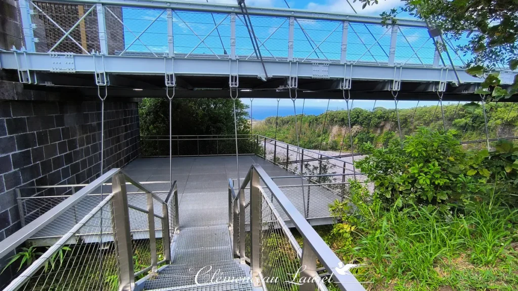 Escalier et passerelle en caillebotis métallique menant à une plateforme d'observation aménagée sous le tablier bleu clair du pont suspendu. Sur la gauche se trouve le soubassement en pierres sombres du monument, et sur la droite, une végétation tropicale dense s'ouvre sur une vue dégagée vers l'océan Indien à l'horizon. - Photo : Clémenceau Lauret (Licence CC BY-SA 4.0)